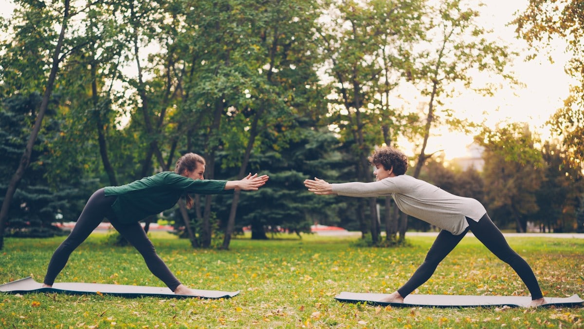 Attractive girls are enjoying outdoor yoga in city park practising positions standing on mats. Instructor is speaking teaching student, both women are relaxed and happy.